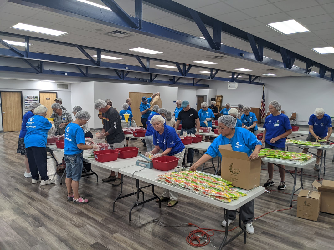 A large, open room with multiple individuals in blue t-shirts and hairnets. They are standing and packing food items on long, white tables.